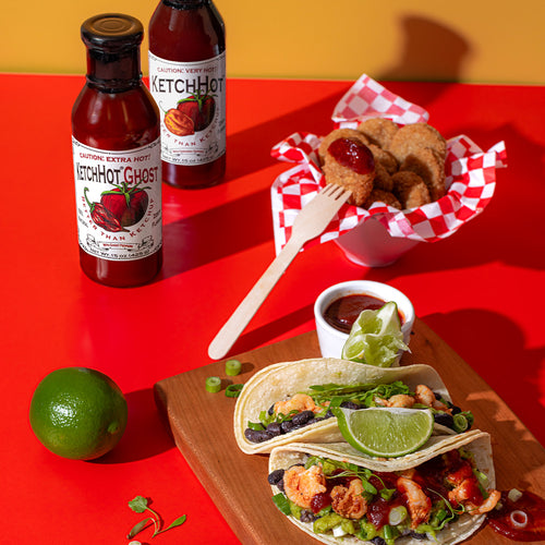 One each of a bottle of KetchHot and KetchHot Ghost on a table next to a lime, tacos, and chicken nuggets